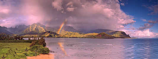 A panoramic fine art landscape photograph of Hanalei, Kauai. A rainbow embellishes the beautiful Hanalei bay and town while the sunset turns clouds a light pink. Mountains in the distance with waterfalls and a passing rain shower make this image unique. Landscape photography by Inspiring Images Hawaii.