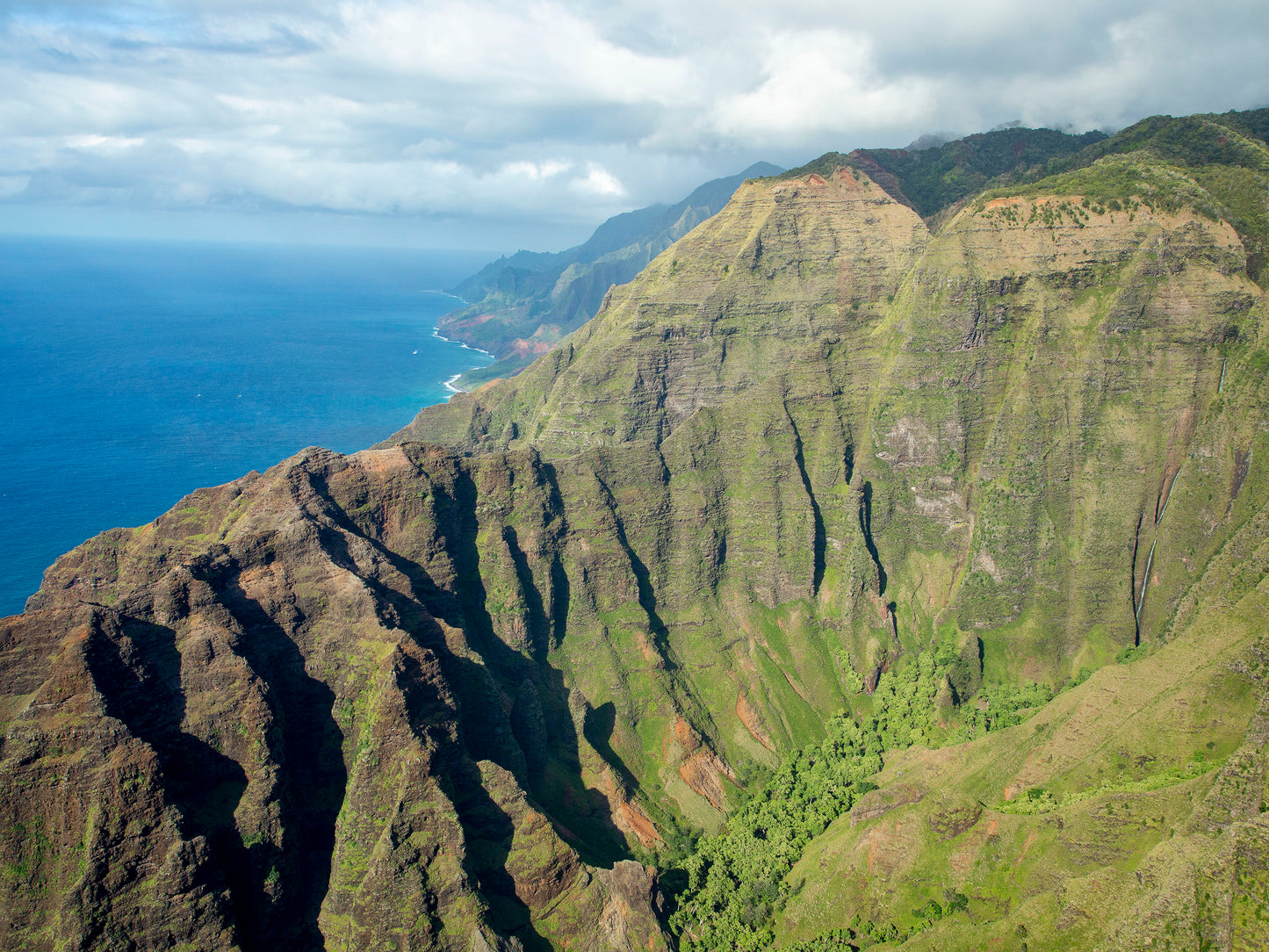 Fine art photograph of Nualolo Valley, one of the valleys along Kauai’s Napali Coast. Steep, green cliffs drop down to the ocean with a waterfall  in the distance. Landscape photograph by Inspiring Images Hawaii.
