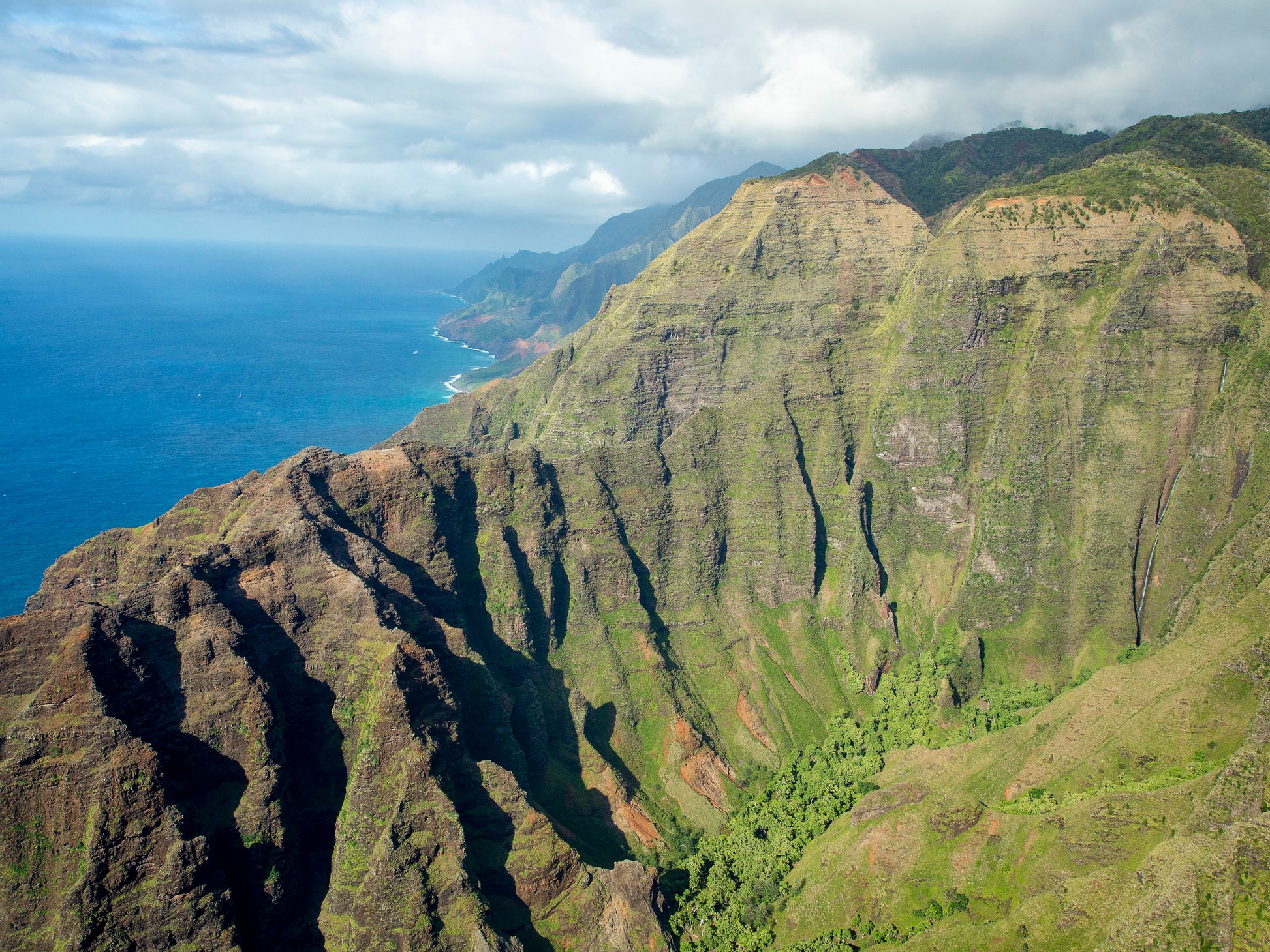 Fine art photograph of Nualolo Valley, one of the valleys along Kauai’s Napali Coast. Steep, green cliffs drop down to the ocean with a waterfall  in the distance. Landscape photograph by Inspiring Images Hawaii.