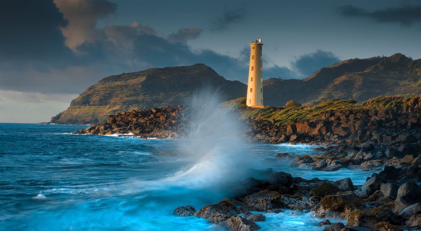 A wave crashes onto the rocks in front of Ninini Point Lighthouse on Kauai. Scenic outdoor landscape photography by Inspiring Images Hawaii, sold as fine art pieces.
