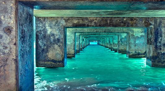 Fine art photograph taken under Kauai’s Hanalei Pier. The bright turquoise water reflects on the rustic concrete underside of the pier as it angles out toward the ocean. Photograph by Inspiring Images Hawaii.