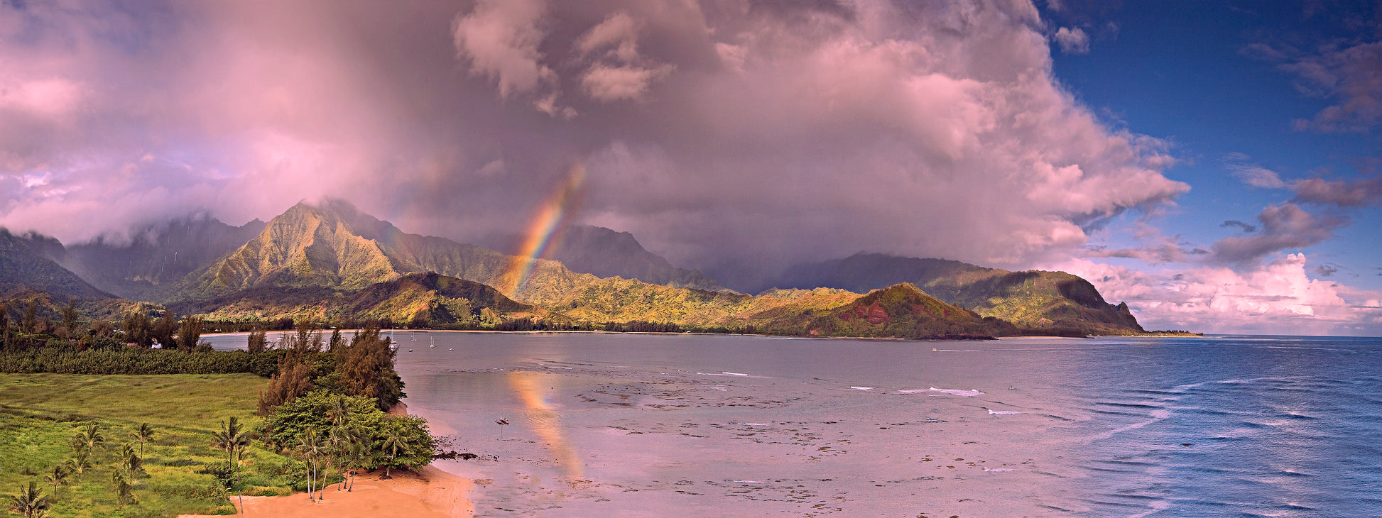 A panoramic fine art landscape photograph of Hanalei, Kauai. A rainbow embellishes the beautiful Hanalei bay and town while the sunset turns clouds a light pink. Mountains in the distance with waterfalls and a passing rain shower make this image unique. Landscape photography by Inspiring Images Hawaii. 