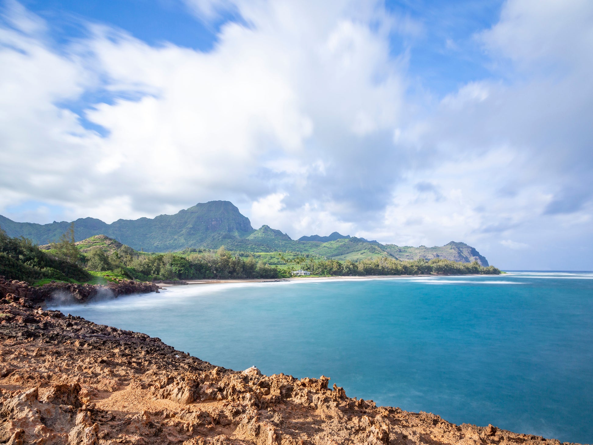 A fine art photograph of Mahaulepu Beach on Kauai. Jagged rocks in the foreground are juxtaposed by soft blue ocean water, the beach, and mountain range in the background. Landscape photograph by Inspiring Images Hawaii.