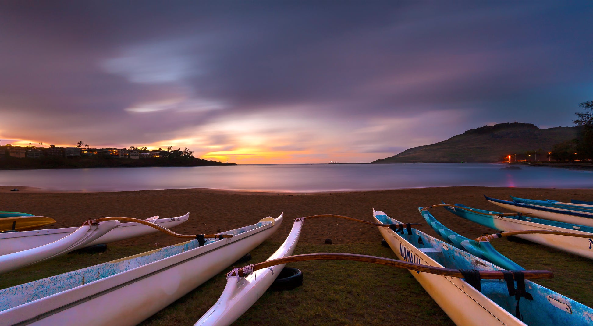 Fine art photograph of outrigger canoes lined up on Kalapaki Beach on Kauai. The photo was taken at sunrise, with a soft purplish blue sky, calm ocean, and canoes with a dim glow from surrounding lights. Landscape photograph by Inspiring Images Hawaii.
