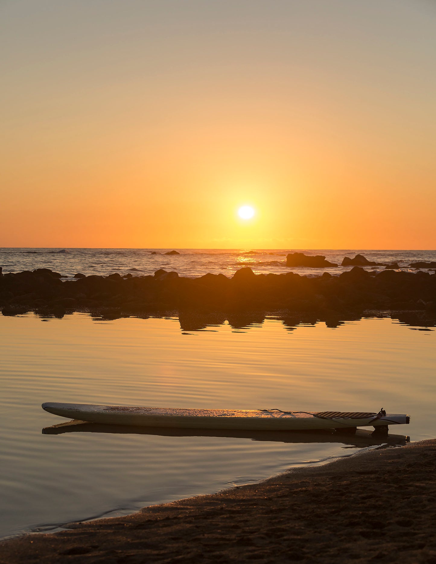A surfboard floats in a calm pool with sunset in background and reflecting on the water. Fine art photograph by Kauai landscape photographers Inspiring Images Hawaii.