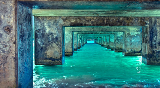 Fine art photograph taken under Kauai’s Hanalei Pier. The bright turquoise water reflects on the rustic concrete underside of the pier as it angles out toward the ocean. Photograph by Inspiring Images Hawaii. 